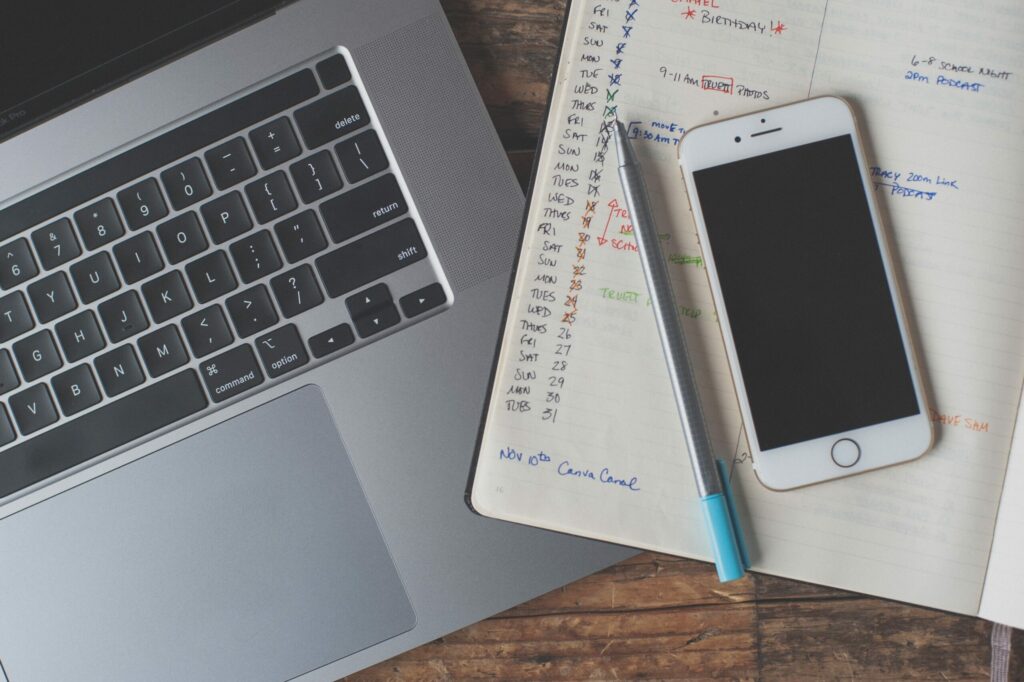 A mobile phone sits on a notebook with a handwritten calendar next to an open laptop.